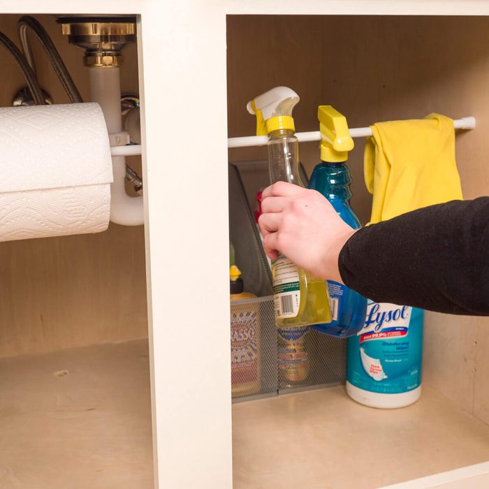 A hand retrieves a cleaning spray bottle from a cabinet, surrounded by other cleaning supplies, paper towels, and a yellow cloth.