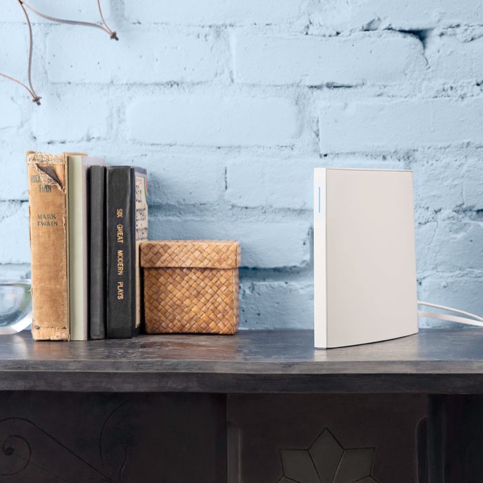 A sleek white object stands on a dark shelf, beside stacked books and a woven container, against a textured light blue brick wall.