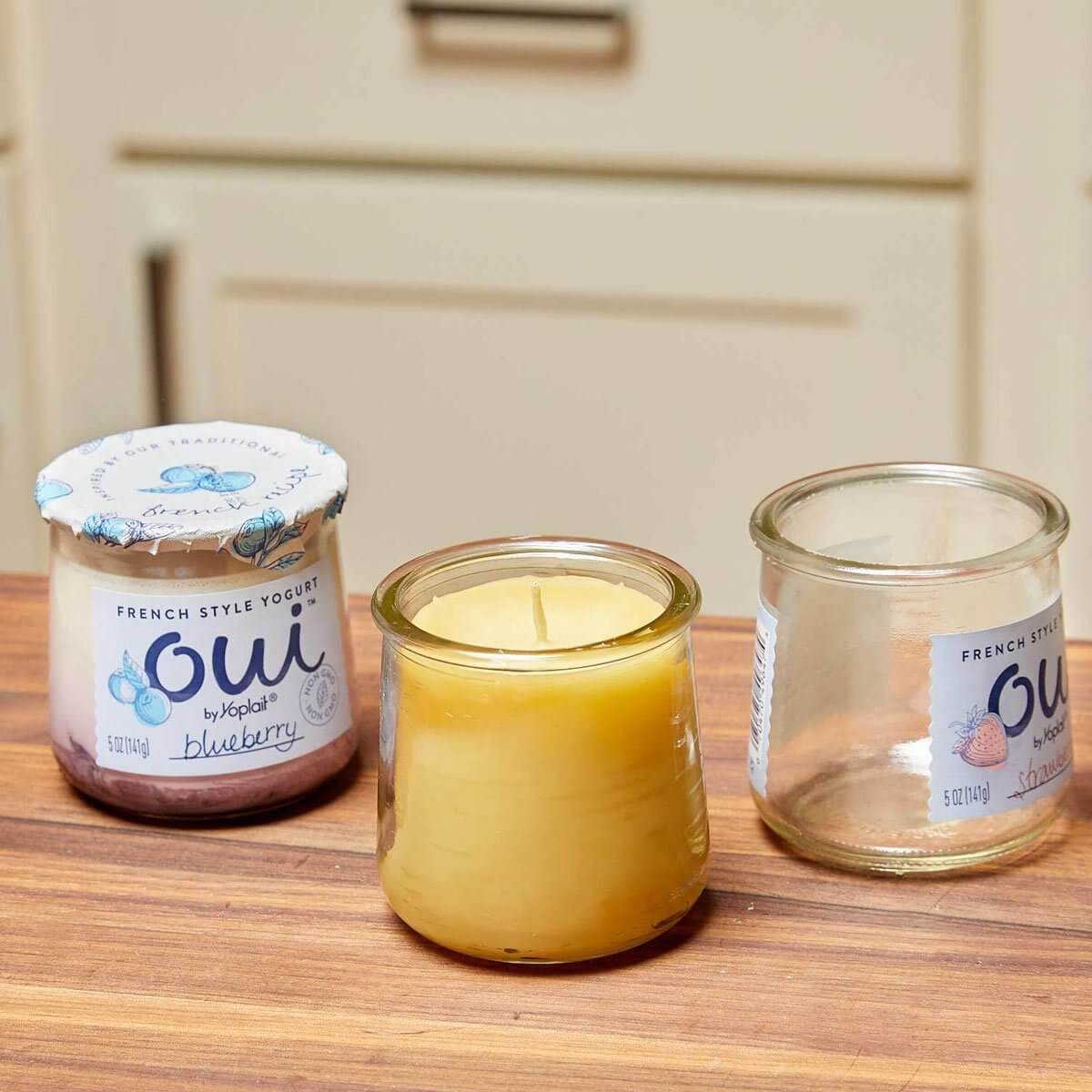 A blueberry yogurt container, a candle, and an empty glass jar are placed on a wooden surface with a kitchen backdrop.