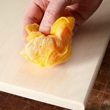 A hand holds a crumpled yellow cloth, applying it to a smooth, light-colored wooden surface in a workshop setting.