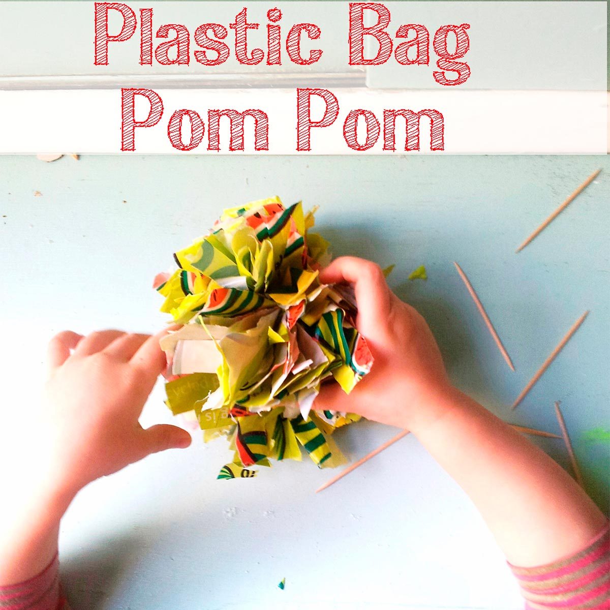 A pair of hands holds a colorful pom pom made from plastic bag scraps on a light blue surface, with toothpicks scattered nearby.