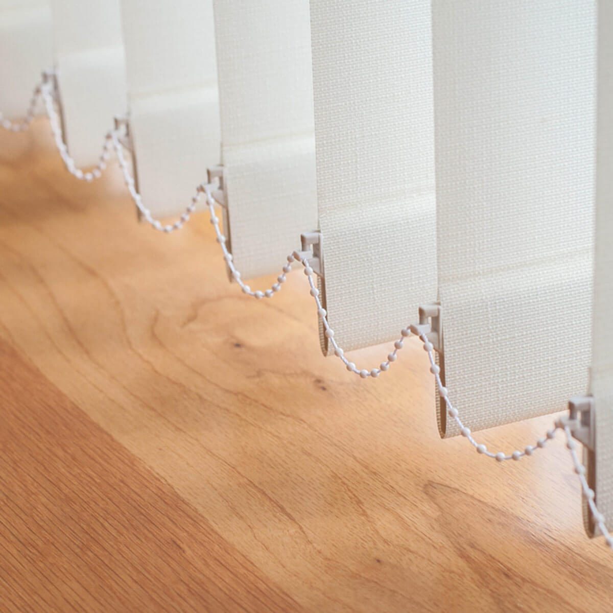 Vertical blinds hang down, gently cascading over a wooden surface, with a white pull chain threaded between them, allowing light control in a bright setting.