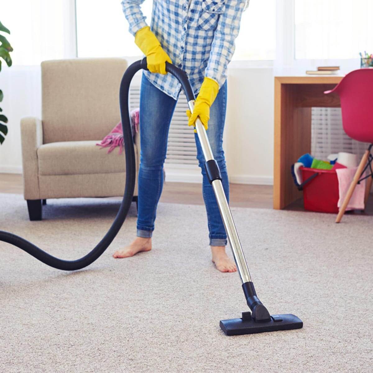 A person wearing yellow gloves vacuums a carpet in a well-lit room, near a sofa and a desk with colorful items.