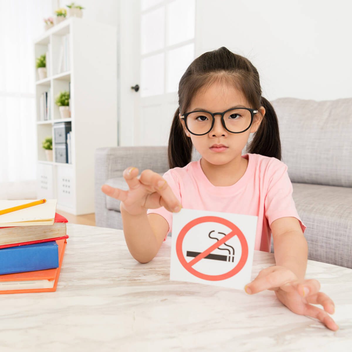 A girl wearing glasses holds up a no-smoking sign, conveying a serious message in a bright, modern living room with books nearby.