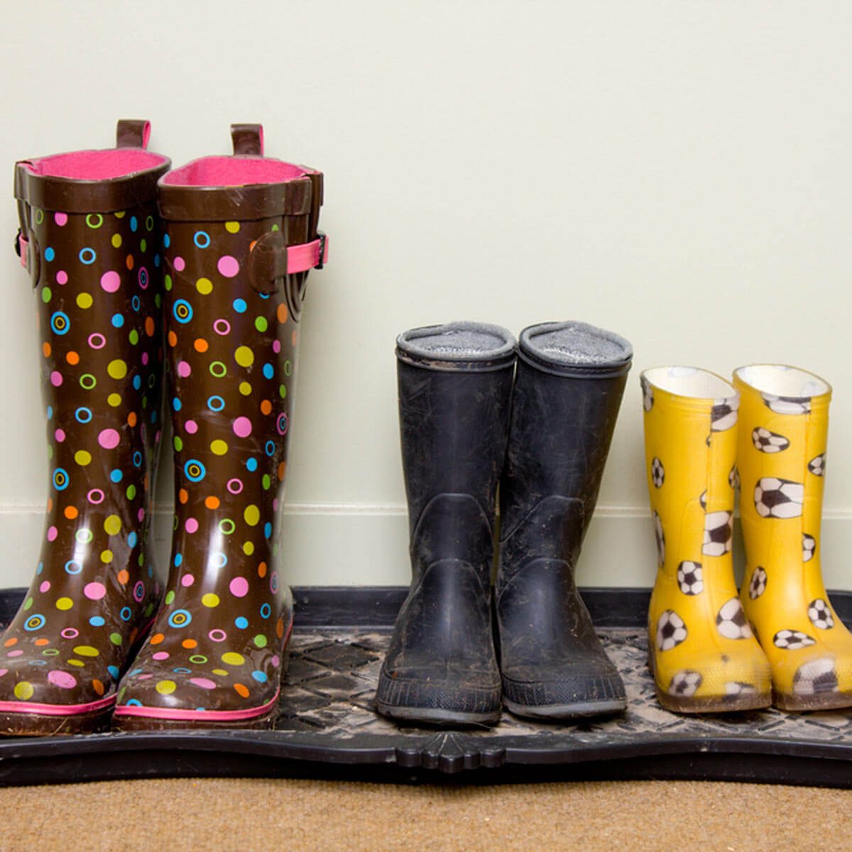 Colorful rain boots stand on a mat, with polka-dotted brown boots on the left, and black and soccer-patterned yellow boots on the right.