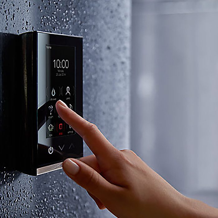 A hand presses a button on a sleek touchscreen control panel mounted on a textured, wet-looking wall, likely in a bathroom setting.