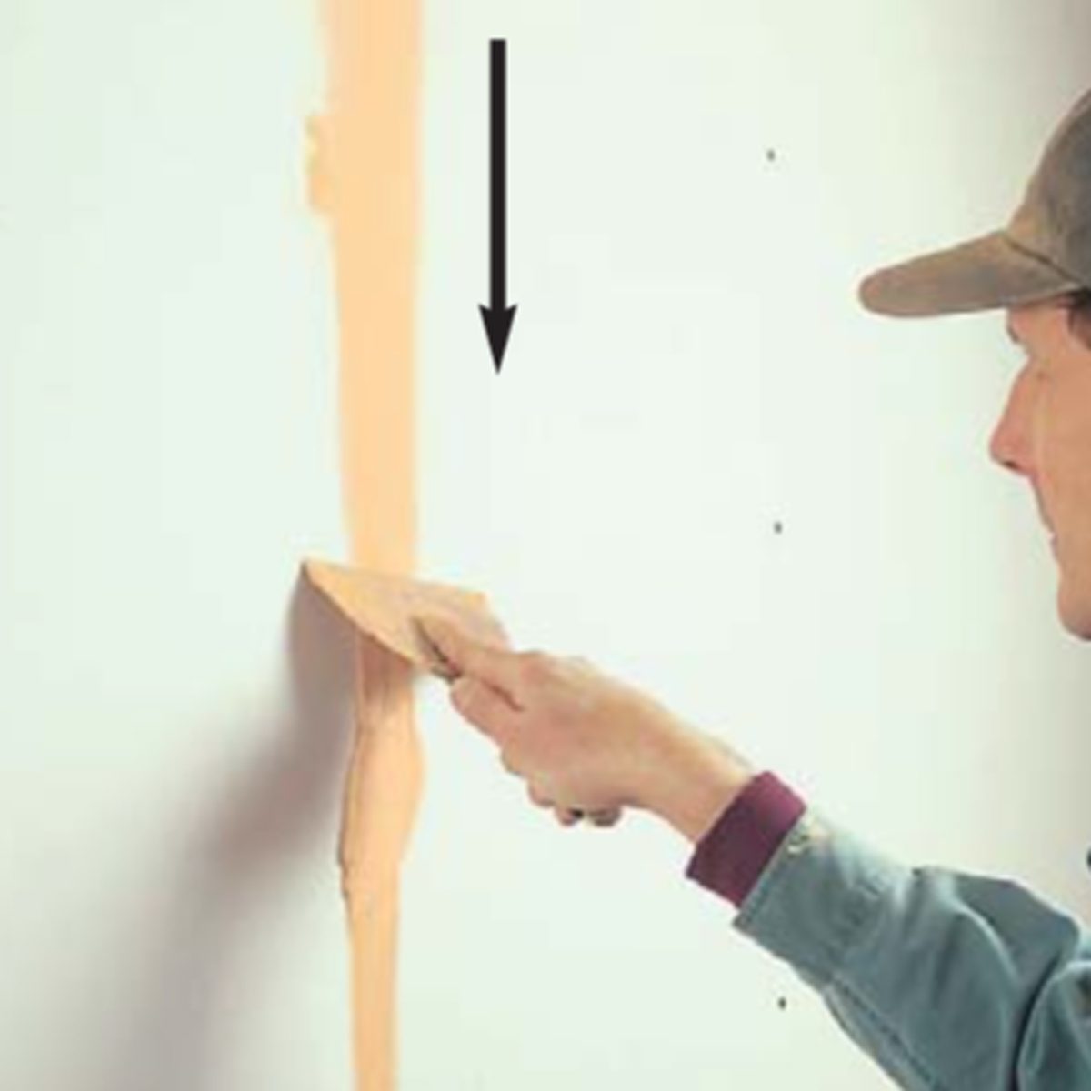 A person scrapes a trowel filled with light-colored substance down a wall, applying it to a visible surface in a plain, indoor setting.
