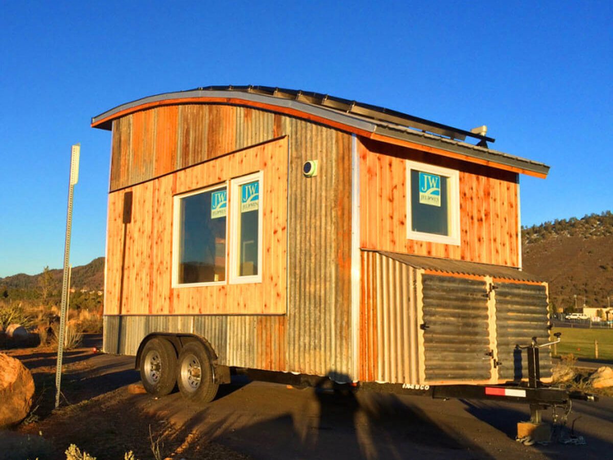 A tiny house on wheels displays a curved roof and mixed wood and metal siding, set against a clear blue sky and rocky terrain.