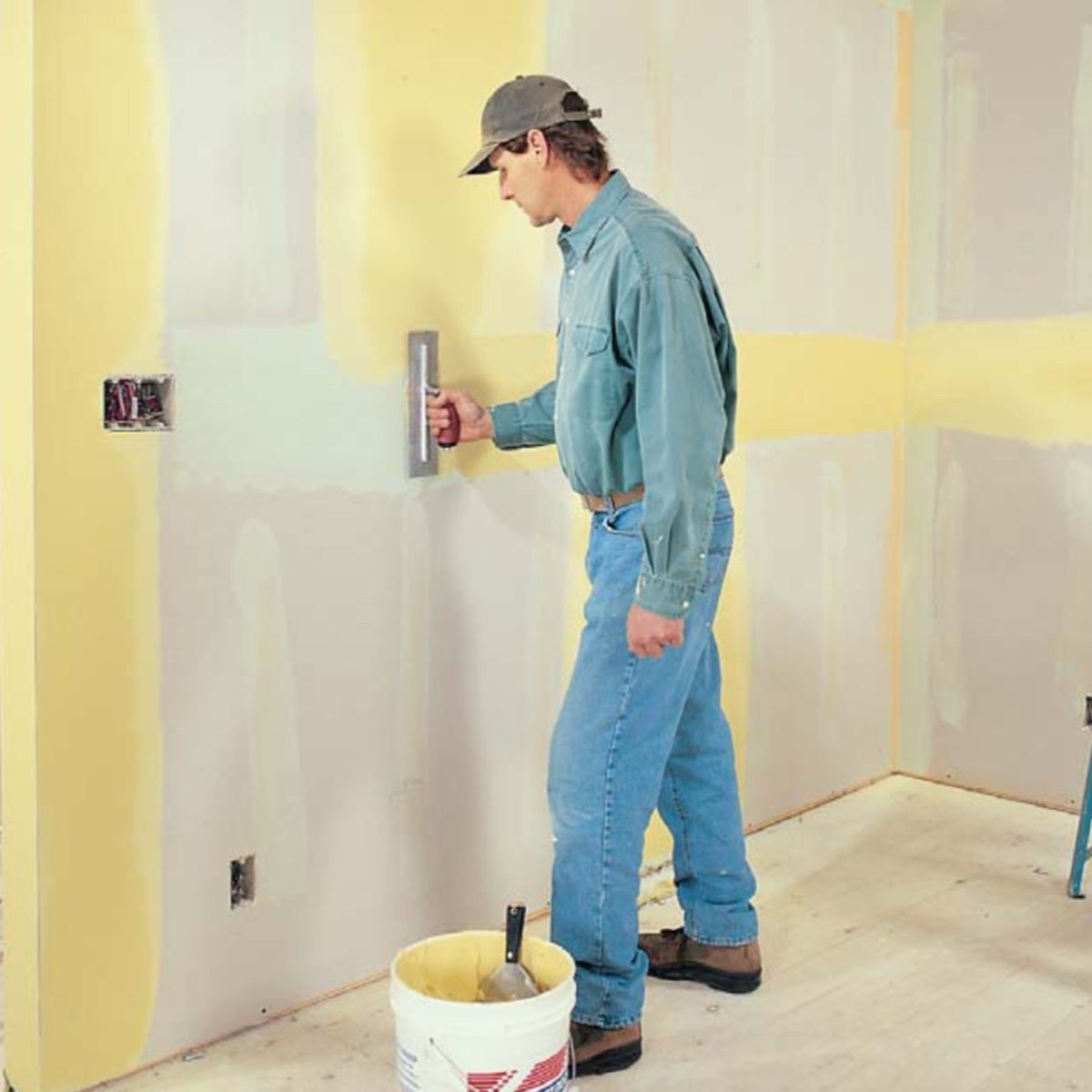 A man uses a smoothing tool on a newly plastered wall in a room with unfinished surfaces and equipment, including a bucket and a ladder nearby.