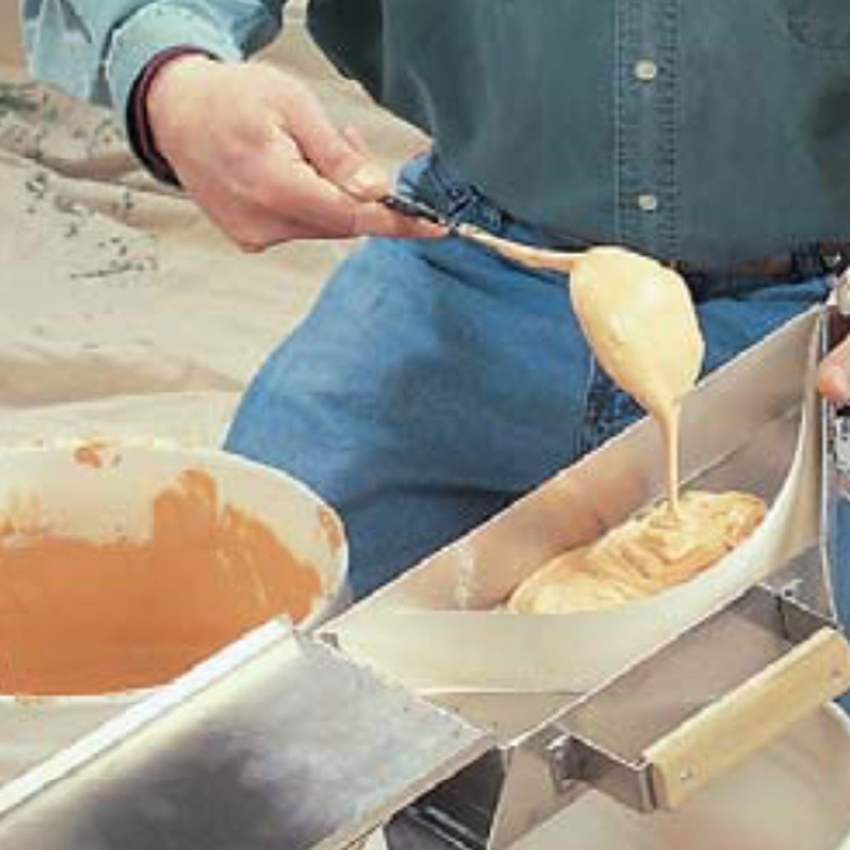 A hand uses a spoon to pour creamy batter into a metal tray, with a bowl of batter placed nearby on a surface.