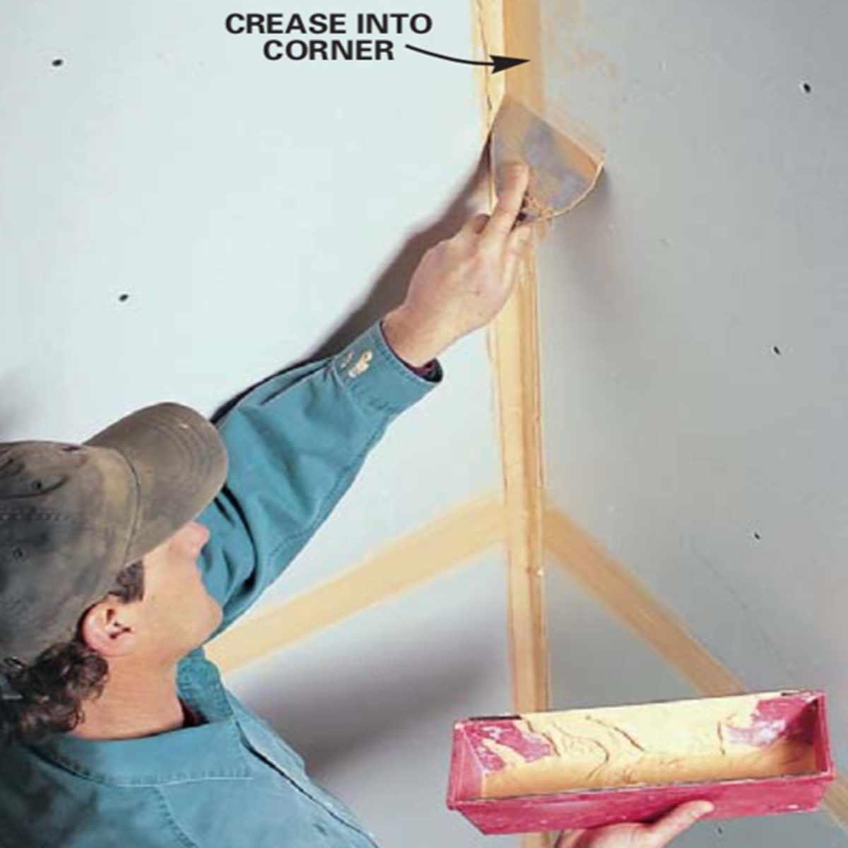 A person applies joint compound into a corner using a trowel, with a tray of the compound in their other hand, against a drywall backdrop.