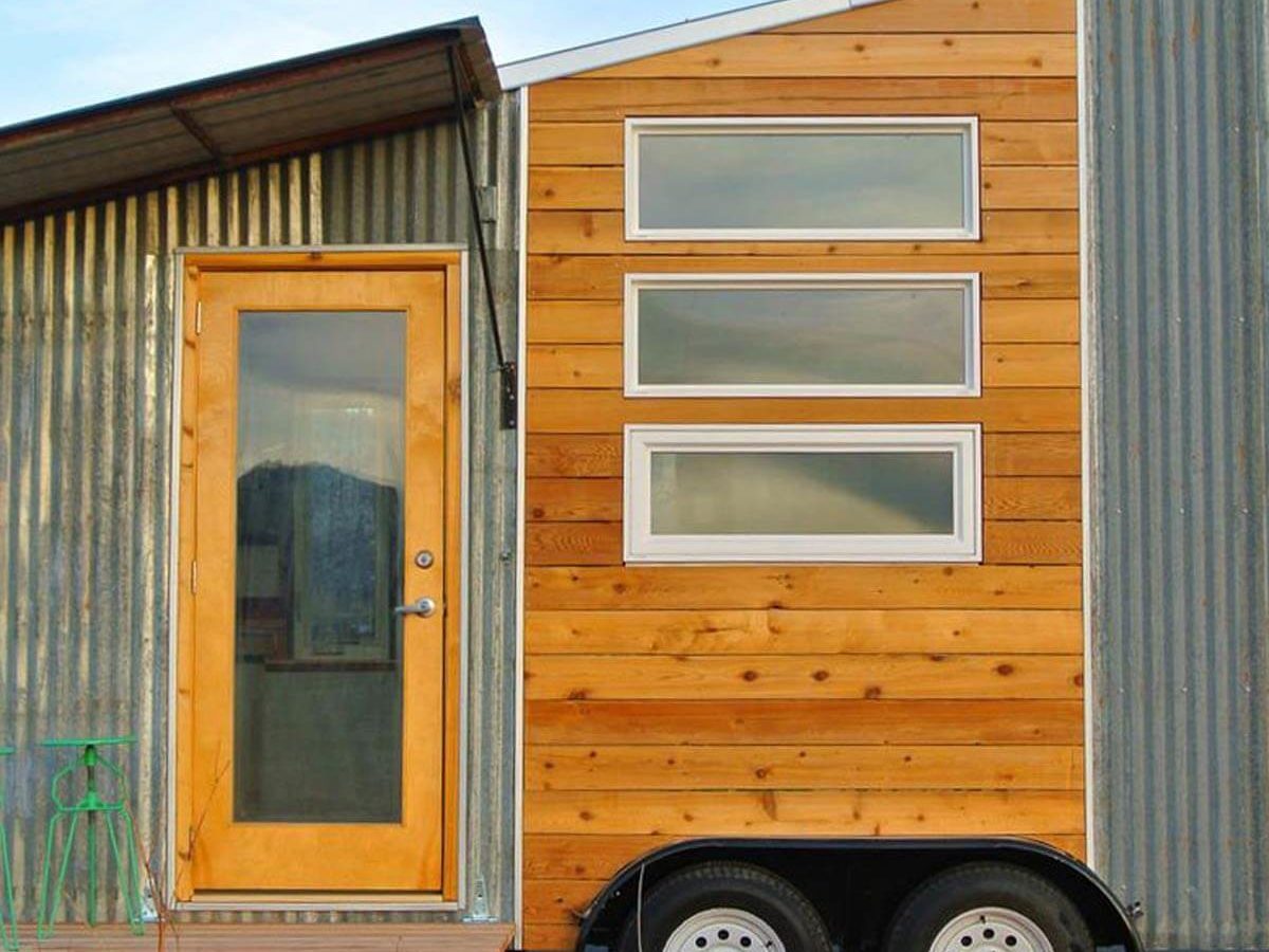 A tiny house on wheels features a wooden door and three windows, surrounded by corrugated metal siding, set against a clear blue sky.