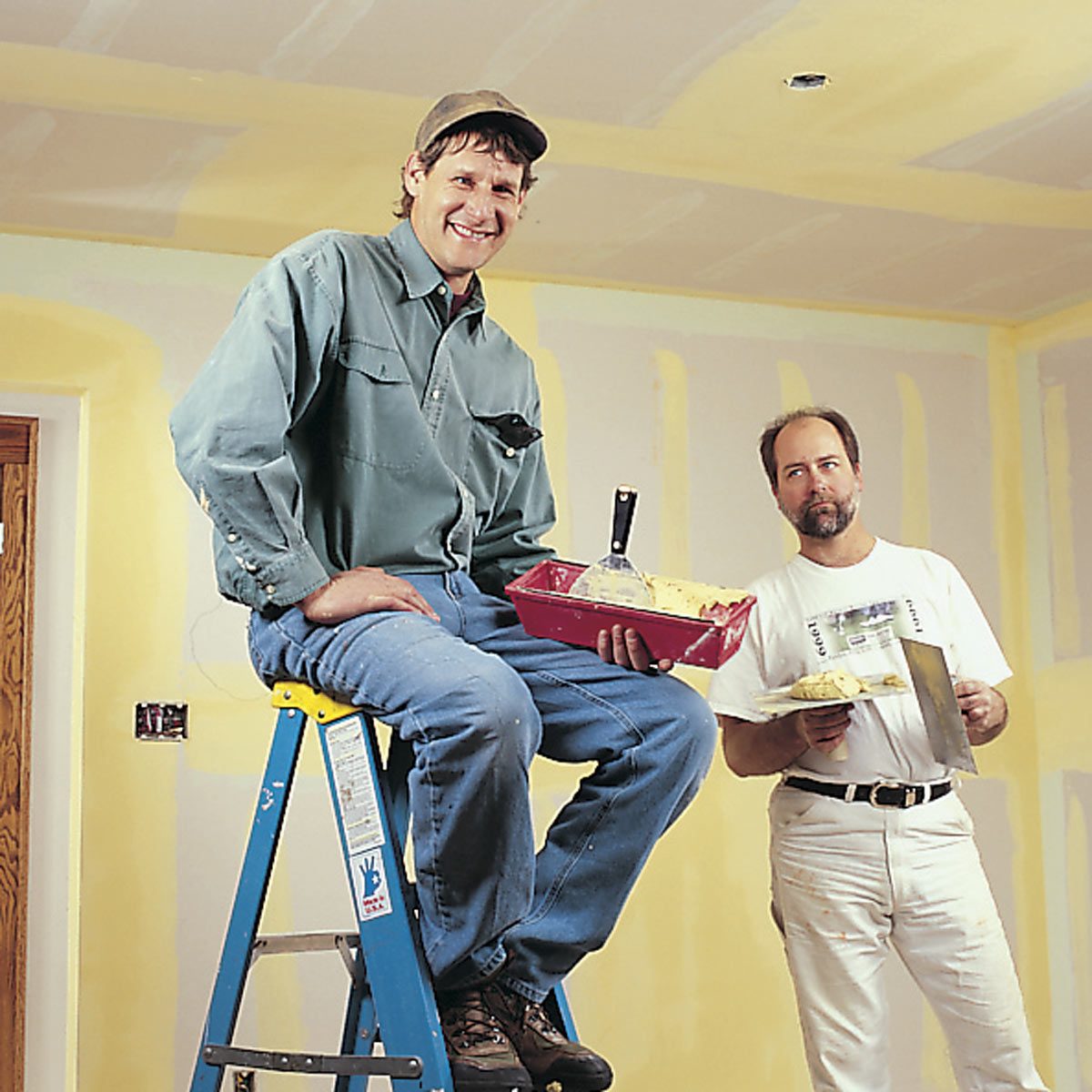 Two men are renovating a room; one sits on a ladder holding a paint tray, while the other stands nearby with a trowel in a newly painted space.