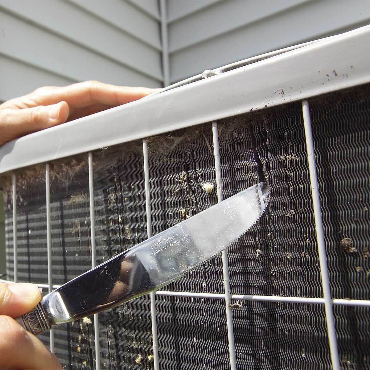 A hand holds a knife, scraping grime from an air conditioning unit's filter. The background shows a house's siding, indicating outdoor maintenance.
