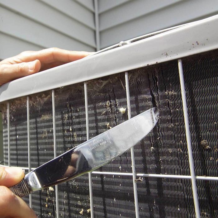 A hand holds a knife, scraping grime from an air conditioning unit