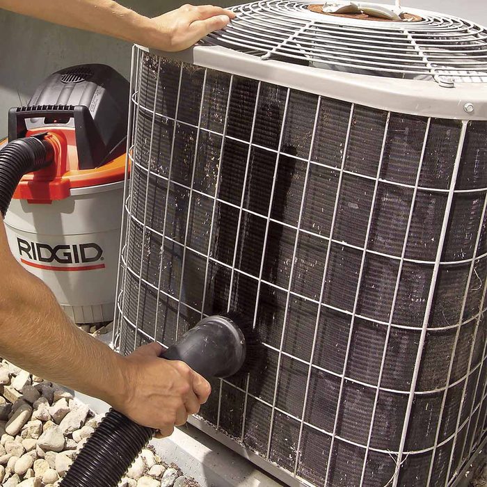A person cleans an air conditioning unit using a vacuum hose, with a RIDGID vacuum nearby, surrounded by gravel and concrete in a sunny outdoor setting.