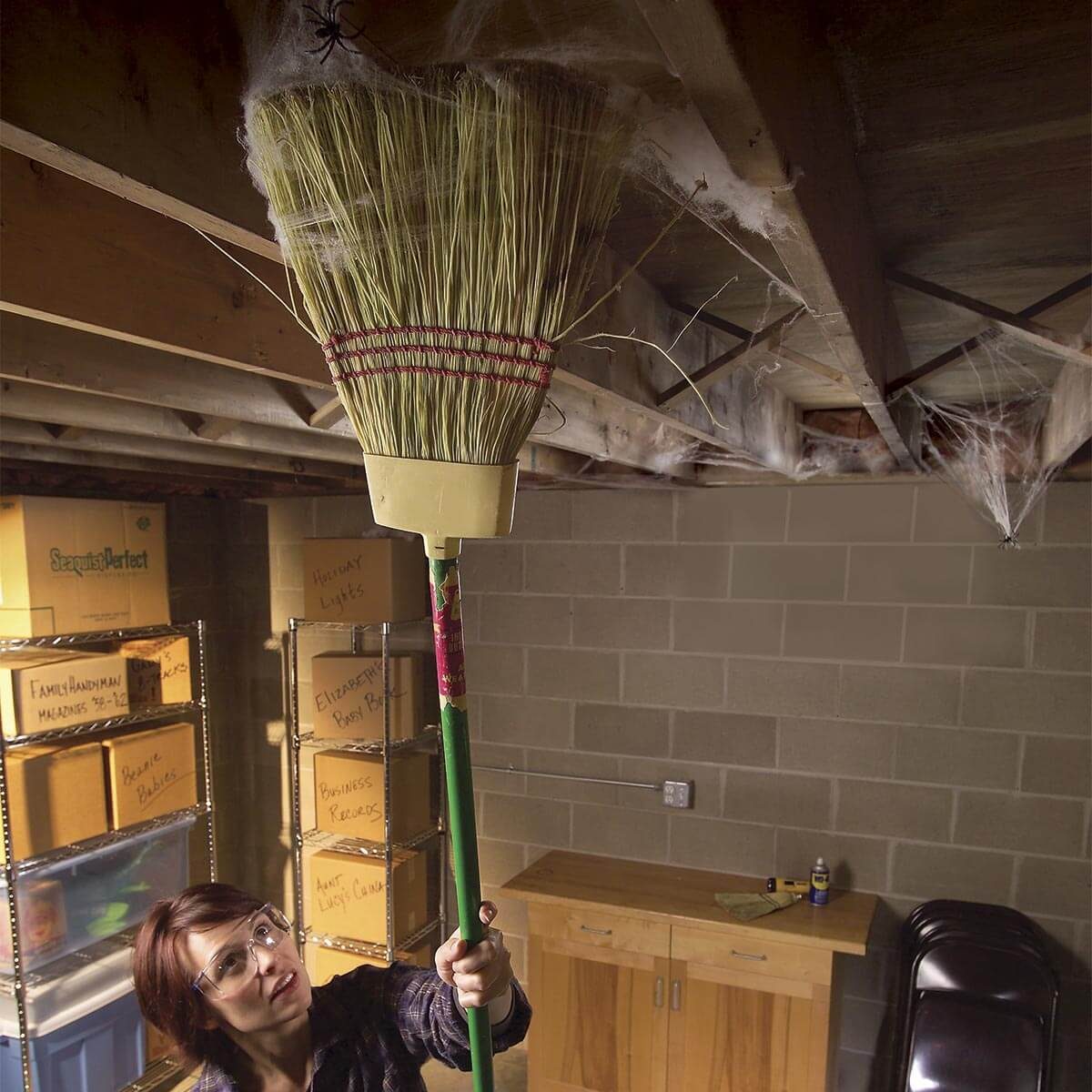 A person uses a broom to remove cobwebs from a basement ceiling, surrounded by storage boxes and shelves in a dimly lit space.