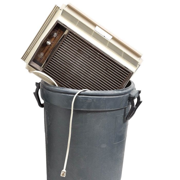 An air conditioner is partially placed in a gray trash can, with its power cord hanging out, suggesting disposal in a cleanup context.