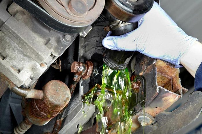 A hand in a glove removes a cap, causing green fluid to spill from a vehicle