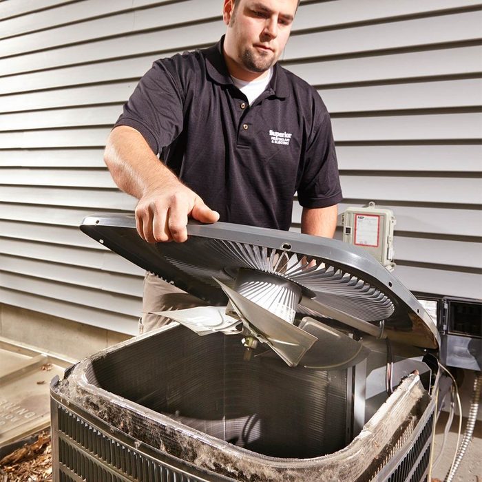 A technician lifts the cover of an air conditioning unit, inspecting its internal components, with a house