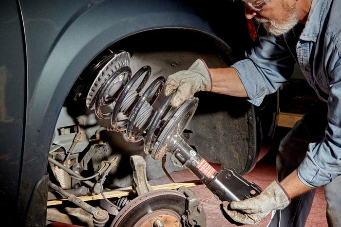 A man, wearing gloves and safety glasses, is removing a suspension coil from a vehicle in a well-lit garage, exposing the brake and suspension components.