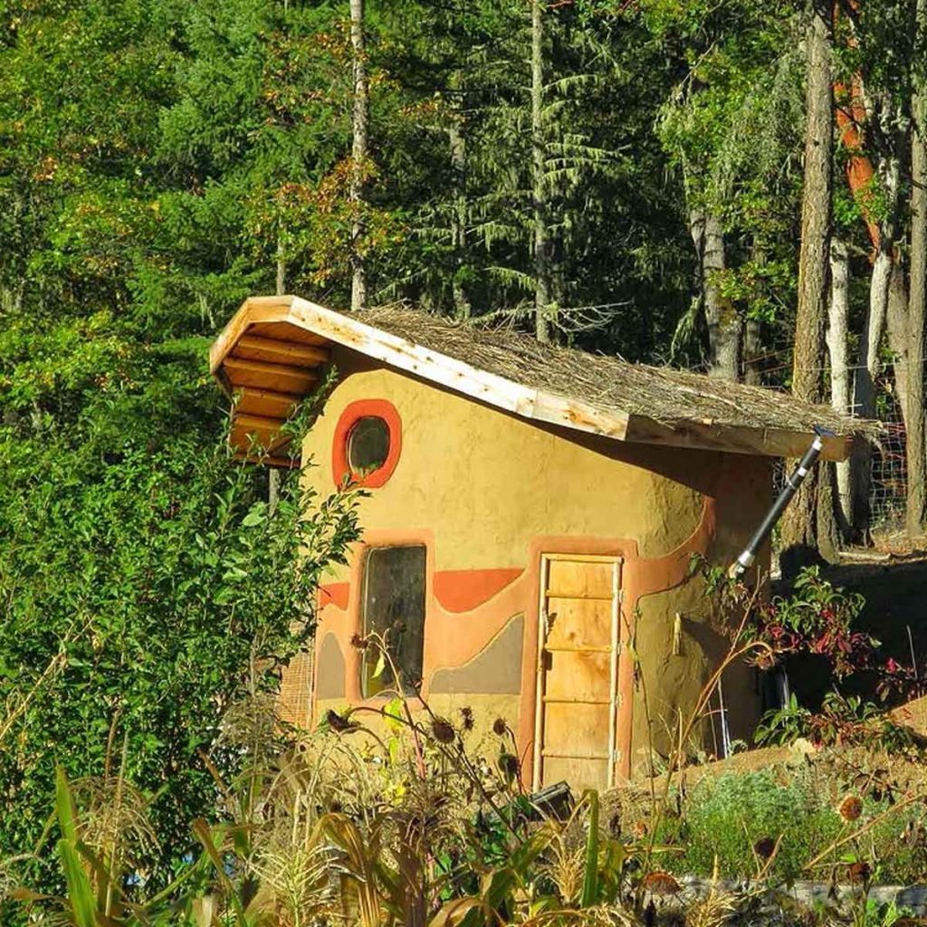 A small, colorful earthen house with a thatched roof sits amidst lush greenery and trees, featuring circular and curved designs on its walls.