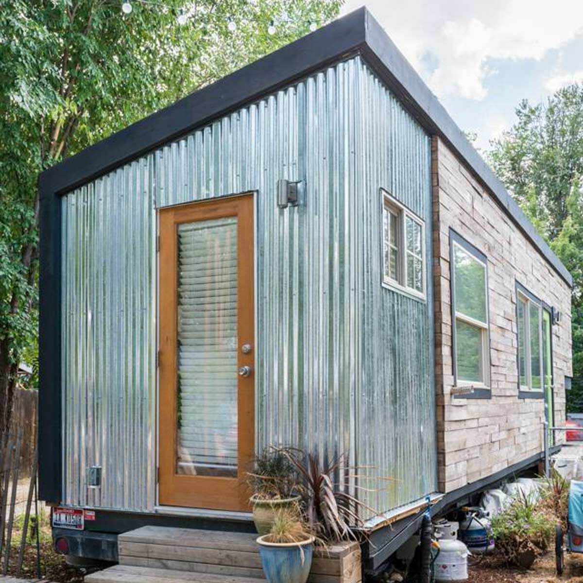 A modern tiny house with metal and wooden siding sits surrounded by greenery. It features large windows, a wooden door, and a small staircase.