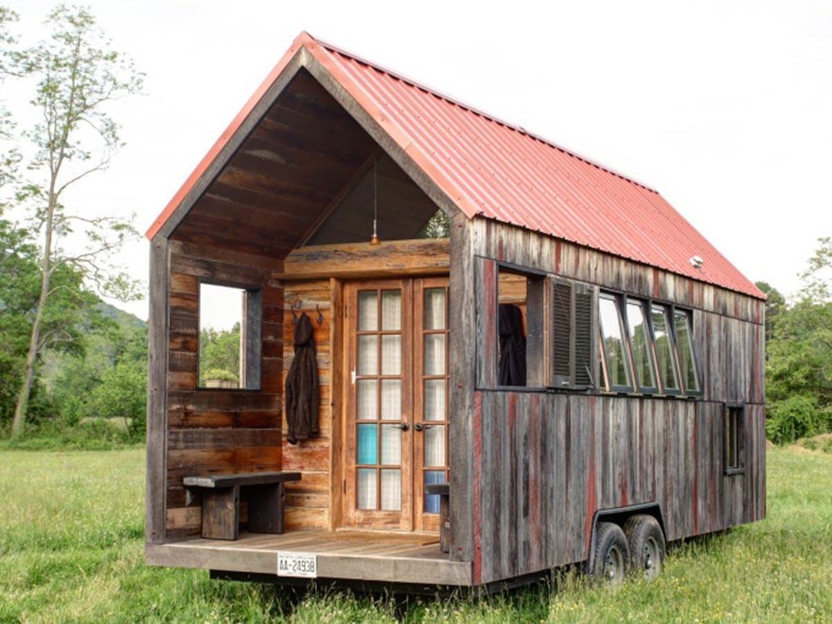 A small, rustic house on wheels sits in a grassy field, featuring a red metal roof, wooden walls, and large windows.