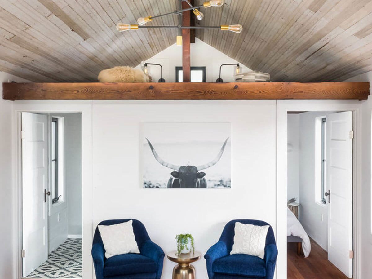A modern living room features two blue chairs facing a coffee table, with a black-and-white bull portrait on the wall and wooden beams above.