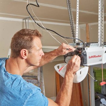 A man repairs a LiftMaster garage door opener, using tools while standing on a ladder in a garage with unfinished walls and visible outdoor access.