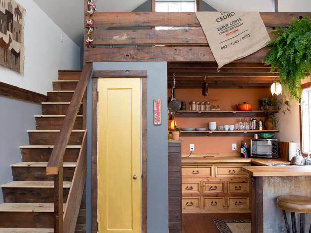 A wooden staircase leads to a lofted area above a cozy kitchen, featuring shelves, a yellow door, and a coffee sack hanging on the railing.