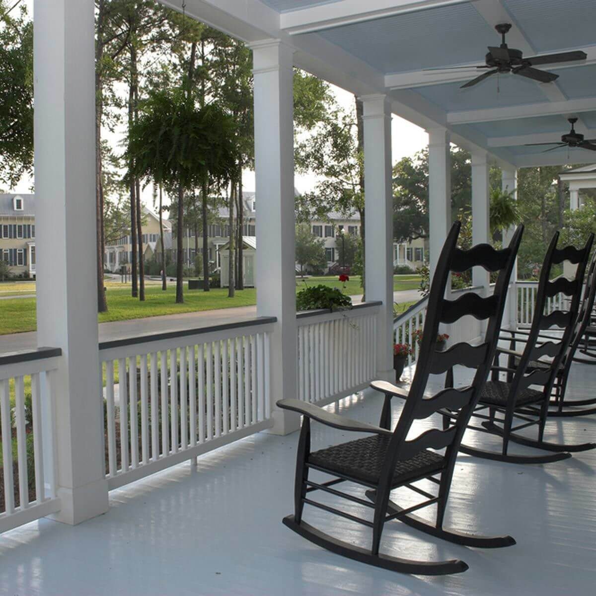 Rocking chairs line a covered porch, gently swaying in a still environment, overlooking a green lawn and nearby trees in a quiet neighborhood.