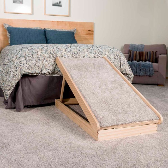 A wooden ramp is positioned next to a bed, assisting pets to ascend onto the mattress, surrounded by a cozy, carpeted room with soft furnishings.