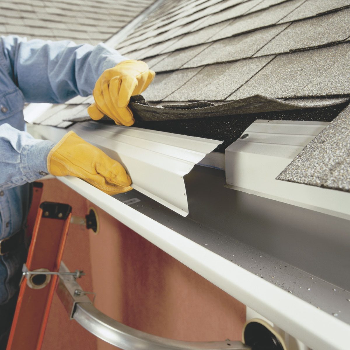 A person in gloves installs a white gutter section, using a tool on a sloped roof with gray shingles, near a ladder against the building.