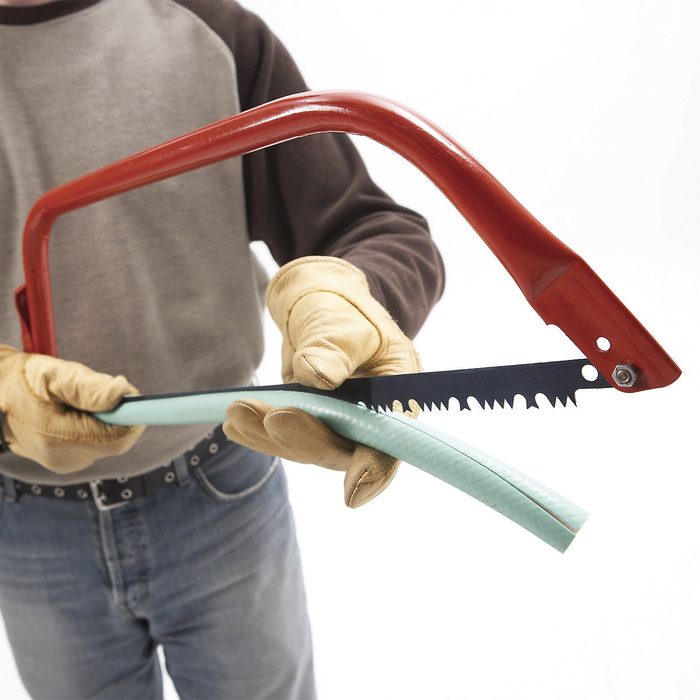 A person wearing gloves holds a handsaw, cutting through a light green hose, with a neutral white background surrounding them.
