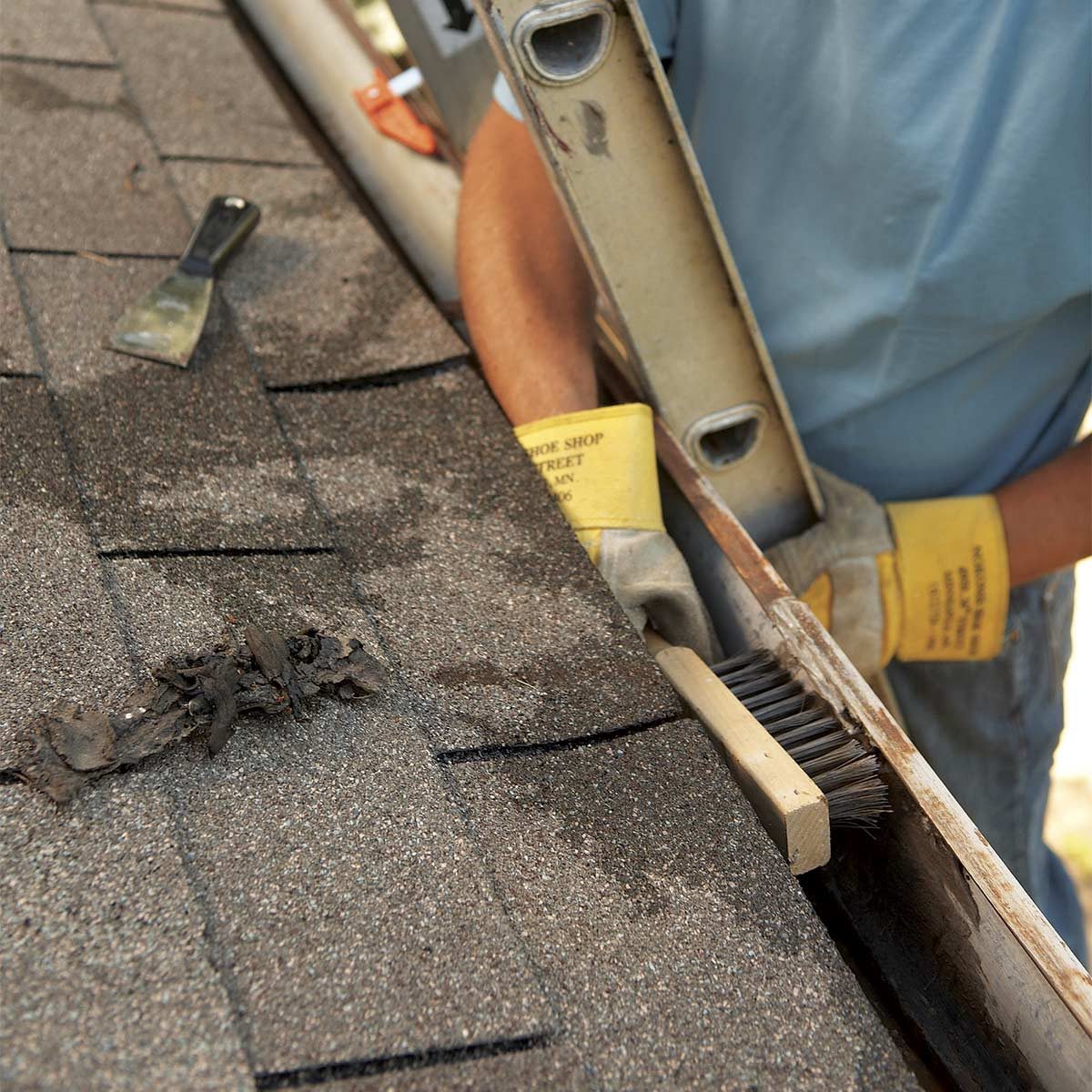 A gloved hand scrapes debris from a roof gutter using a brush, while a ladder leans against a structure under clear skies.