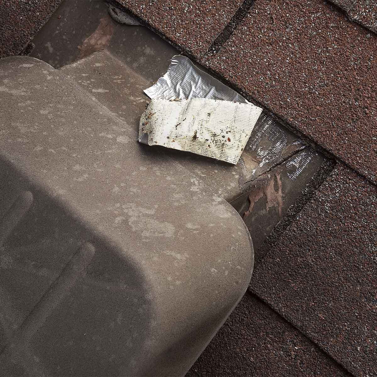 A weathered roof vent cap sits on shingles, partially covered by a torn piece of metallic tape, indicating repair or patchwork in a residential setting.