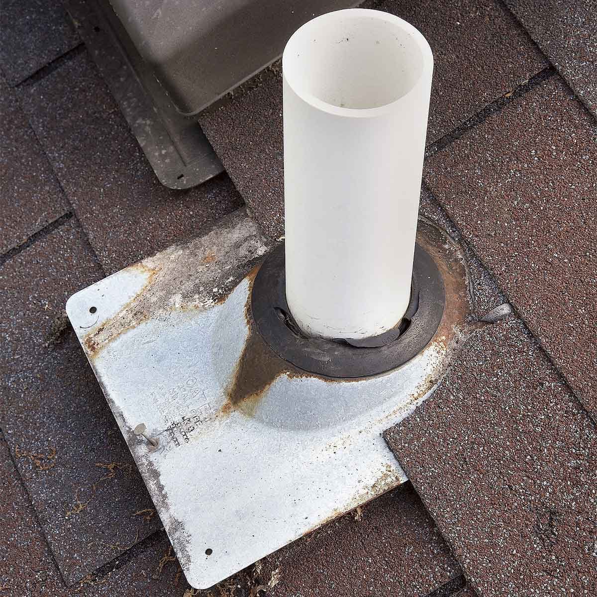 A white pipe rises from a metal flange, mounted on weathered roofing shingles, displaying signs of rust and wear in a residential setting.