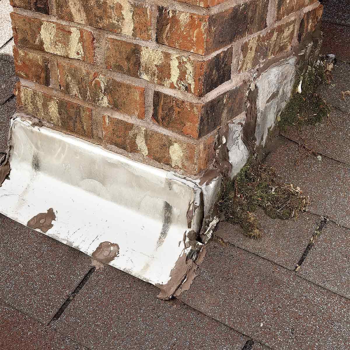 A brick chimney base is partially covered with weathered metal flashing, showing signs of wear and some moss growing on the surrounding roof surface.