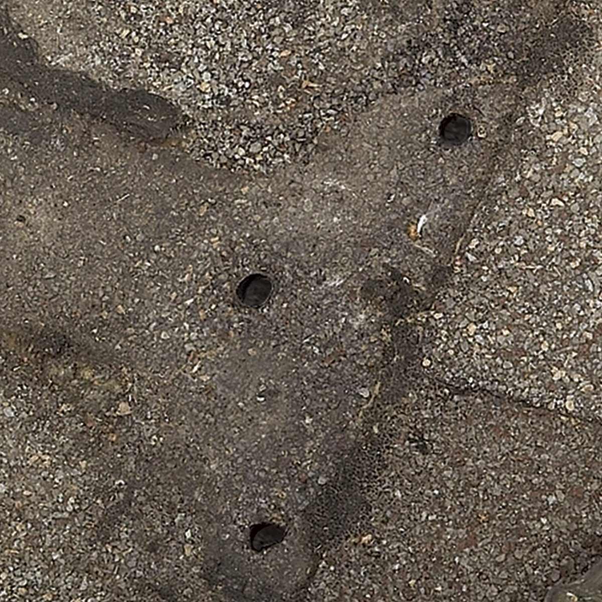 A textured, uneven gray surface features three circular holes; small stones and debris are present in the surrounding area, suggesting outdoor pavement wear.