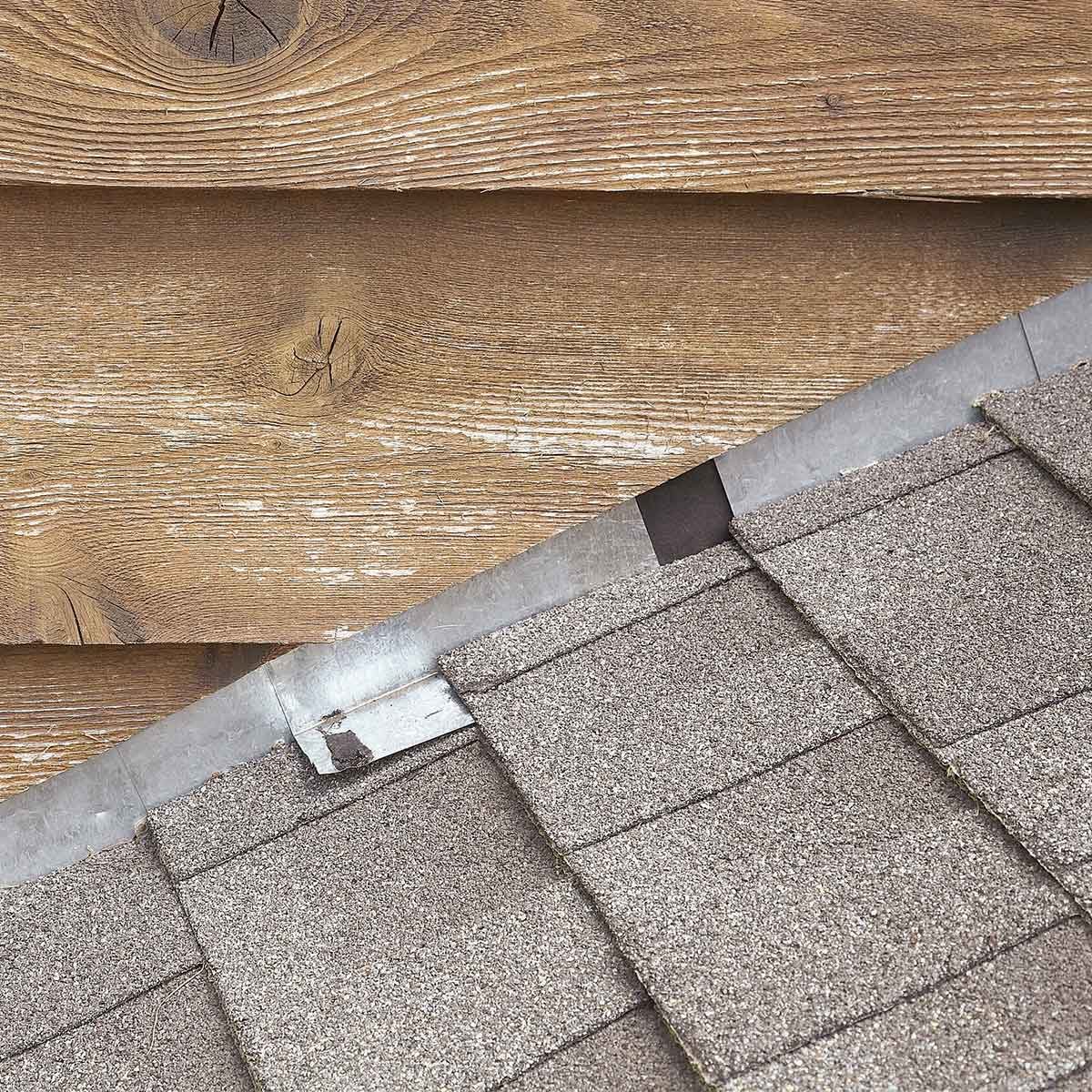 A wooden wall meets a sloped, asphalt shingle roof. The shingles are layered, with metal flashing visible where they connect to the wall.
