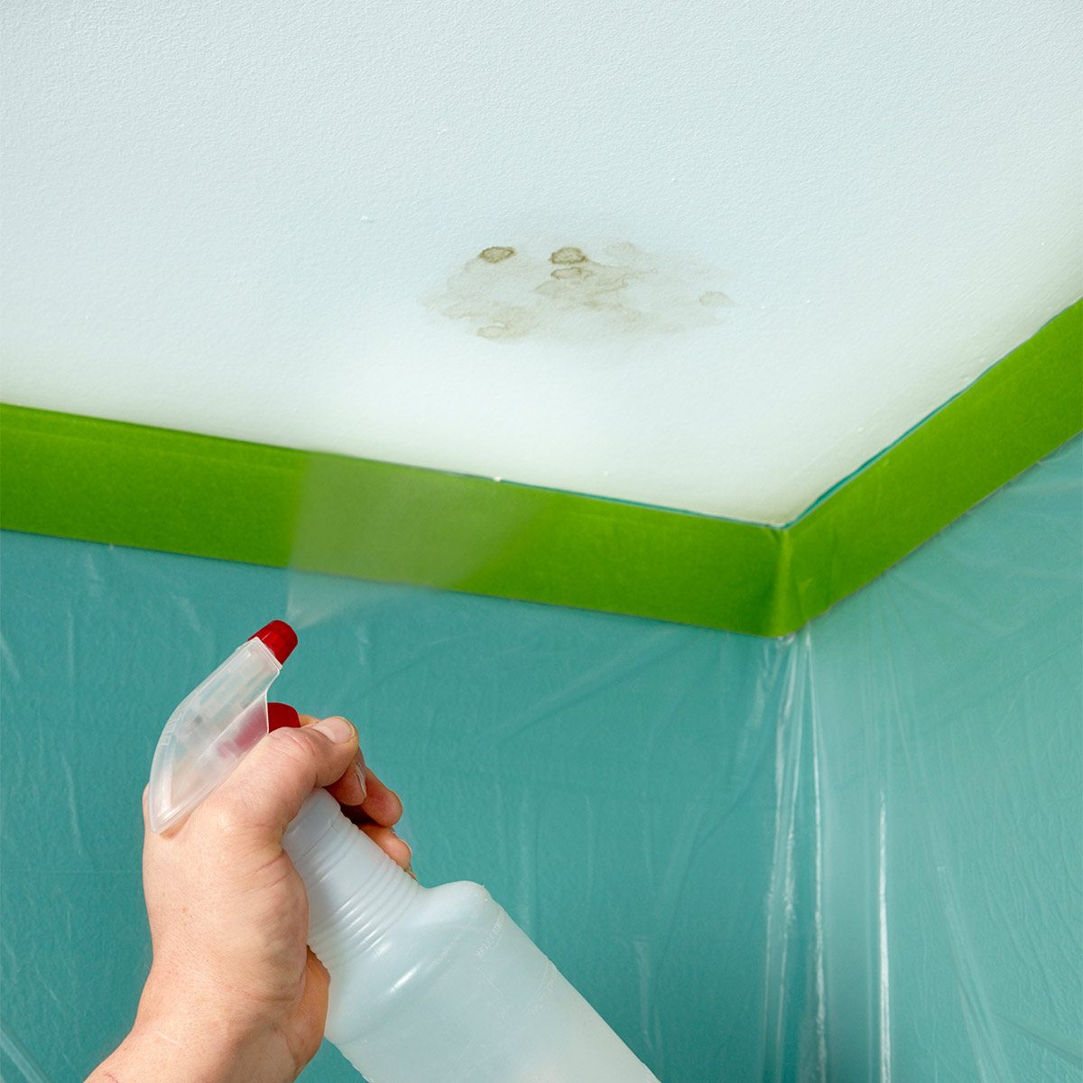 A hand holds a spray bottle, misting a stained ceiling's corner, surrounded by green painter's tape and protective plastic sheeting on light blue walls.
