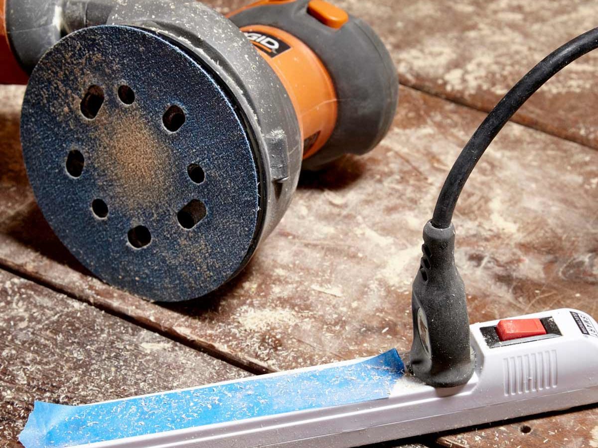 A sander lies on a wooden surface, surrounded by dust, connected to a power strip with blue painter's tape, indicating an ongoing renovation or woodworking task.