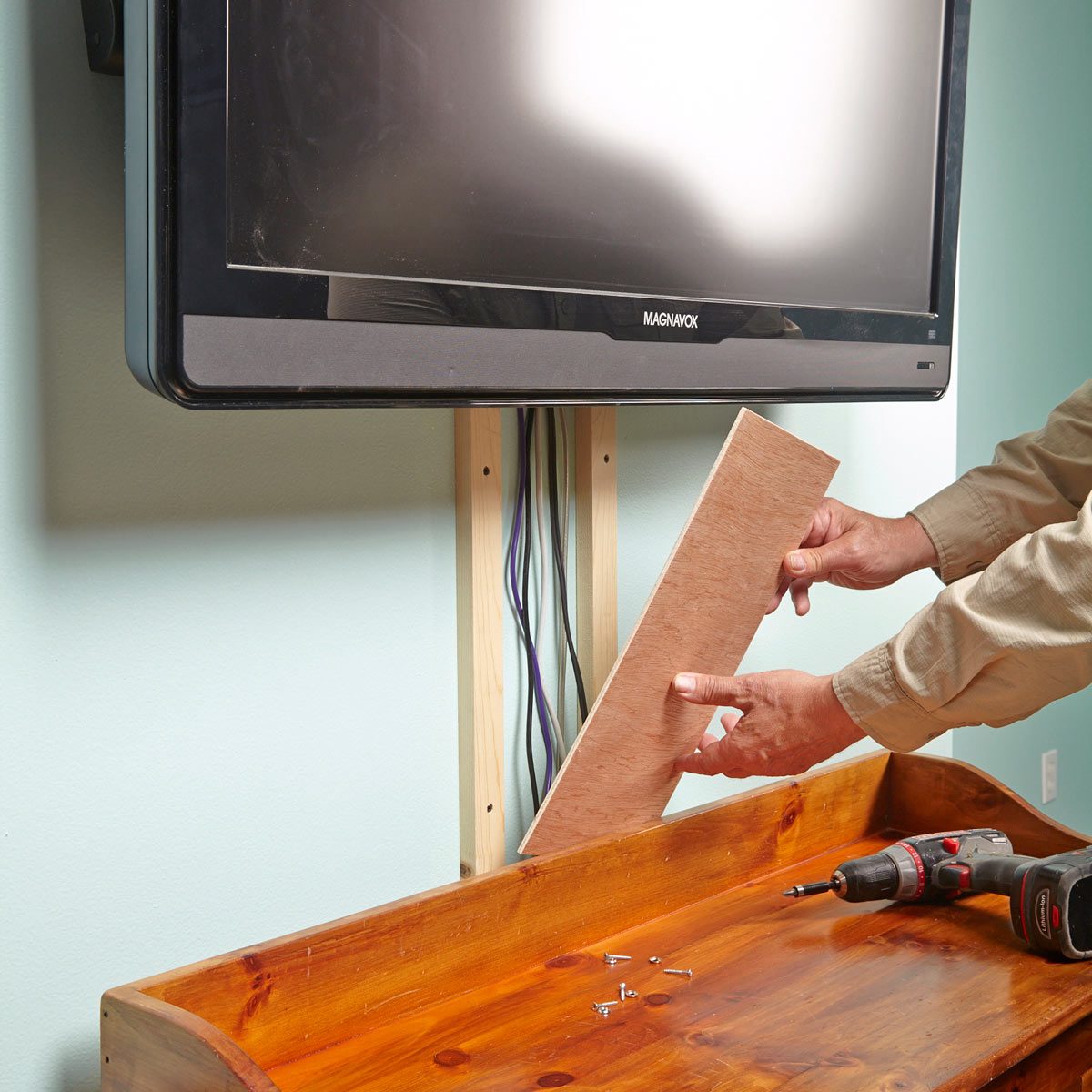 A person holds a wooden panel while installing it behind a mounted TV, surrounded by tools and a wooden shelf on a light-colored wall.