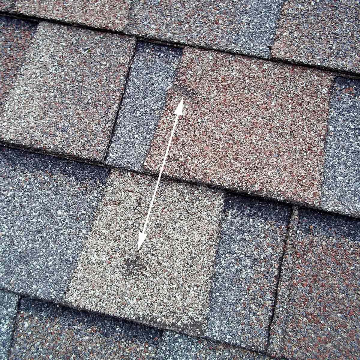 A section of asphalt shingles lies on a roof, showing a missing piece and surrounding intact tiles, highlighting wear or damage in the roofing material.