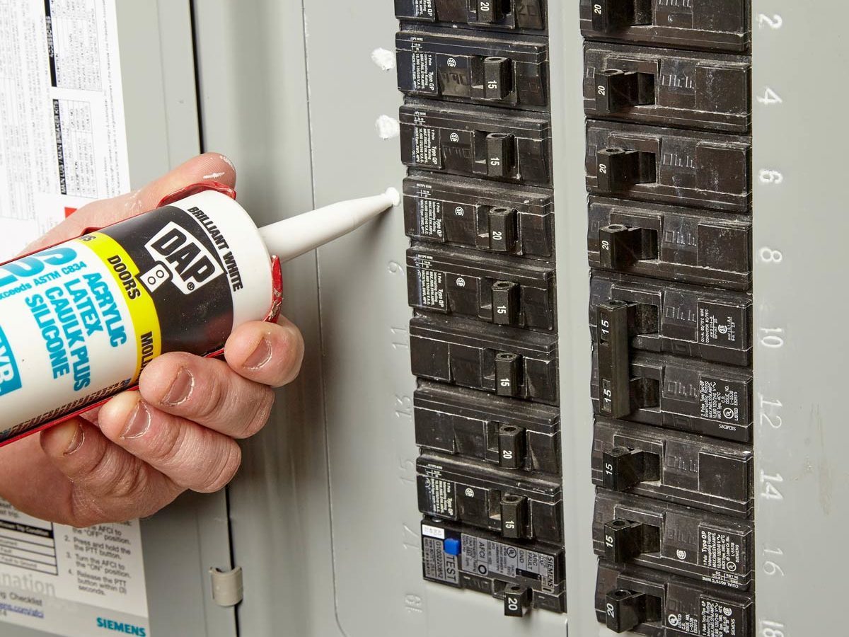 A person applies caulk from a tube to a circuit breaker panel, which displays multiple labeled switches, in a home environment.