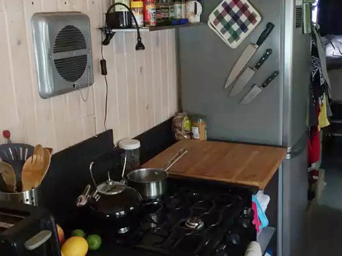 A kitchen features a stove, kettle, and wooden cutting board, with a fridge and utensils nearby, set against a wooden wall.