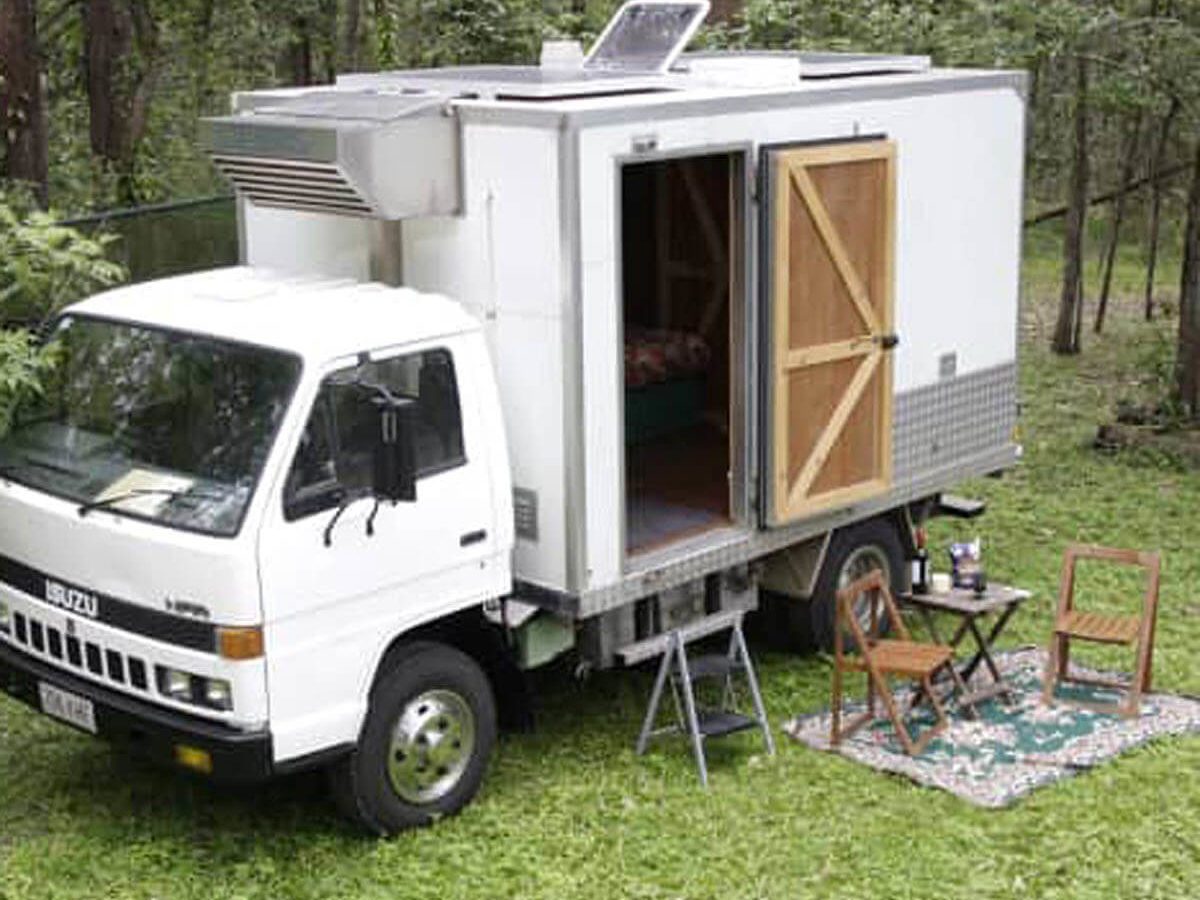 A white truck with an open side reveals a small living space, situated on grass surrounded by trees, with outdoor furniture set up nearby.