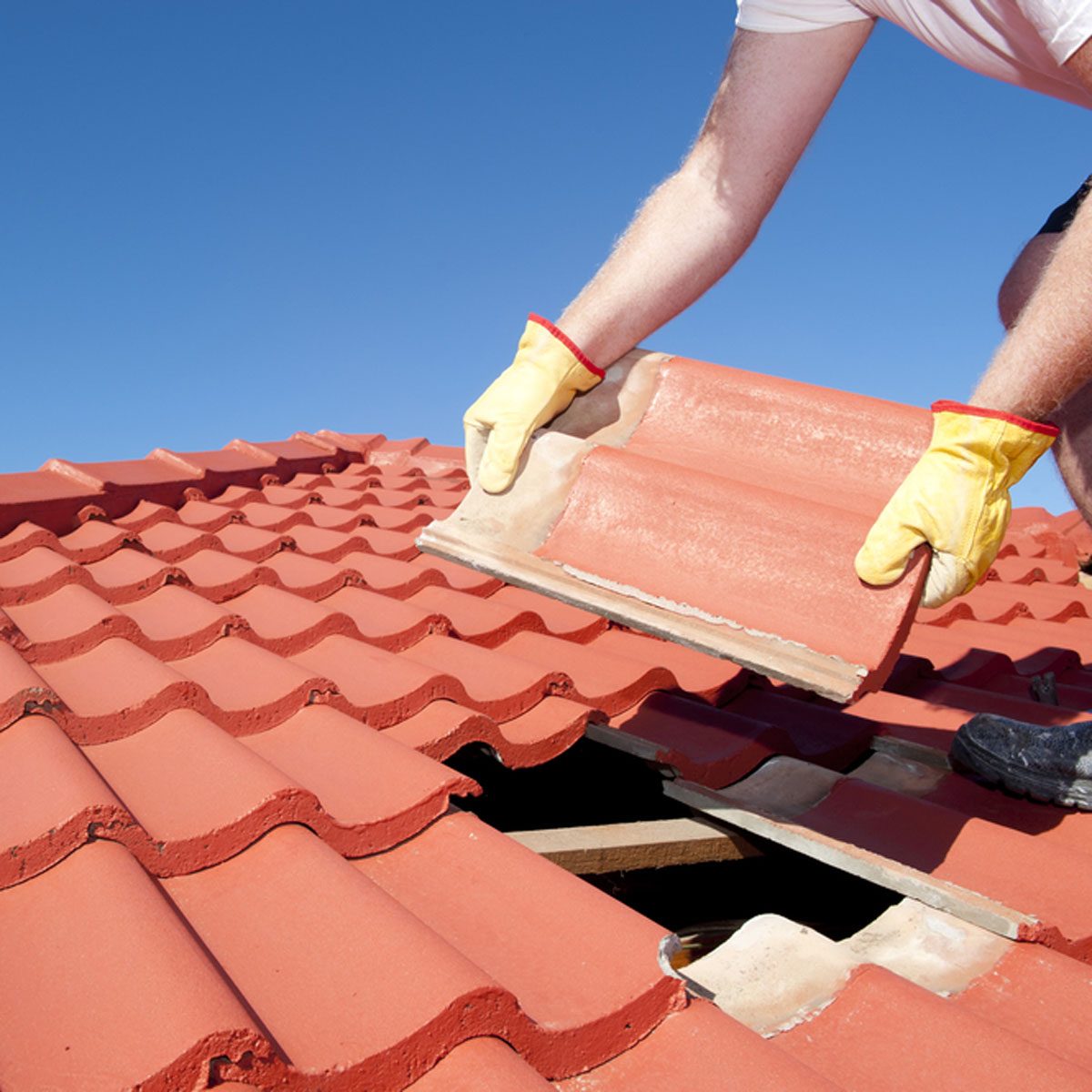 A person in gloves lifts a clay roof tile to replace it, working on a roof under a clear blue sky, with a gap visible between tiles.