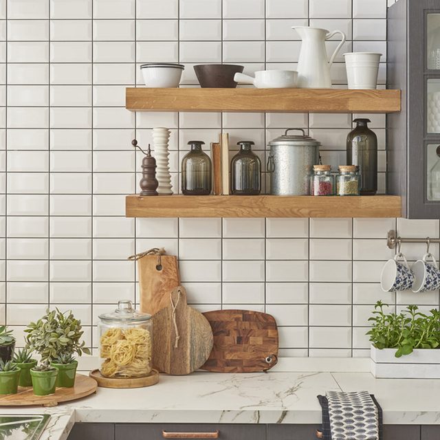 Shelves display various kitchen items, including jars and bowls, while a marble countertop holds plants and cutting boards, all against a tiled white wall.